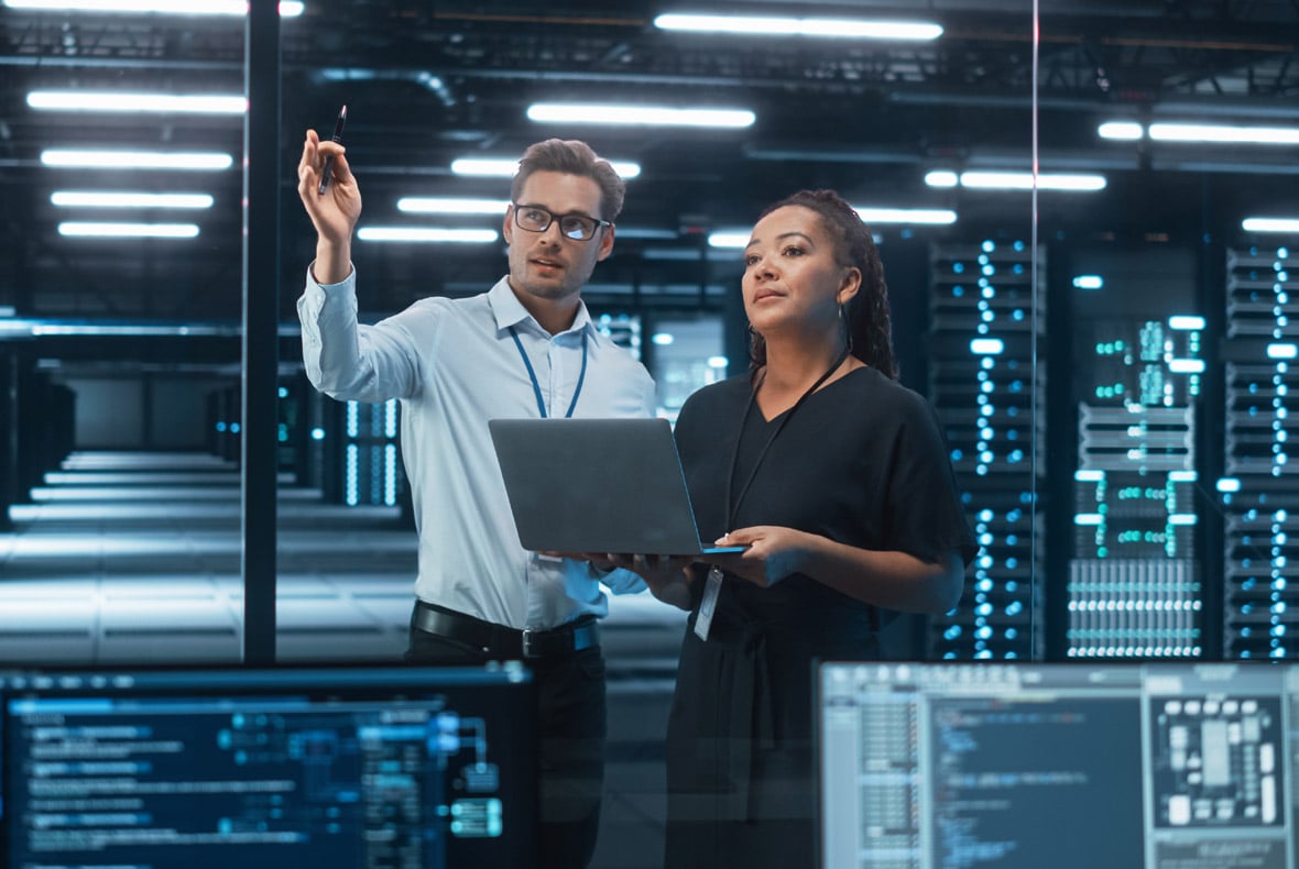 Two IT professionals in a data center discuss information on a laptop, with a man pointing upward and server racks with blue indicator lights visible in the background.