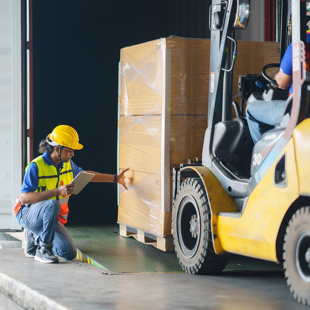 A person in hardhat and safety vest inspects a pallet being loaded by a forklift